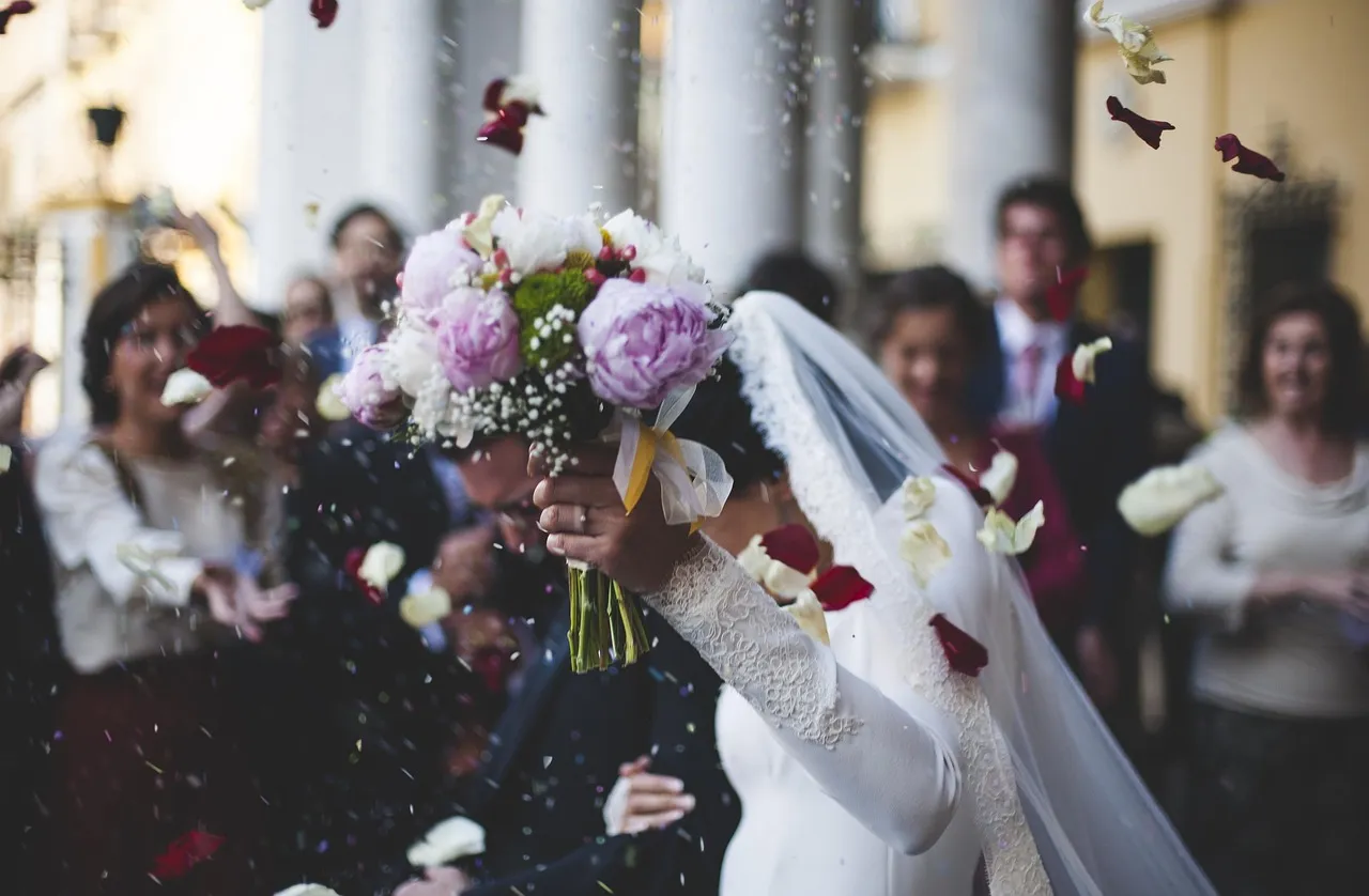 Bride holding a peony bouquet under falling rose petals during a wedding celebration managed by Peter Rowland.