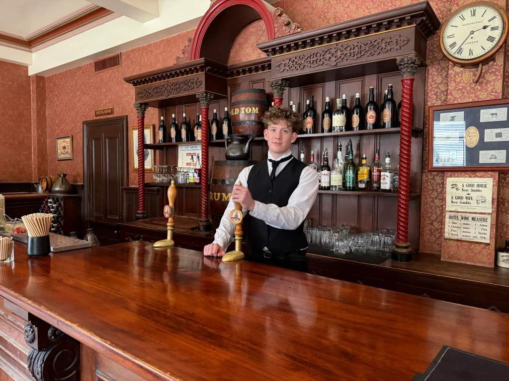 Bartender in Victorian-era period attire standing behind an authentic polished wooden saloon bar at a Sovereign Hill event space.