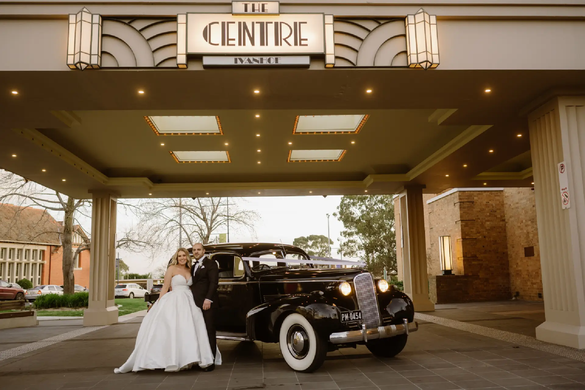 Bride and groom posing with a vintage black car under the entrance canopy at The Centre Ivanhoe.